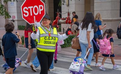 Crossing Guard in neon green vest holds a stop sign, in the background children cross the street carrying and pulling backpacks